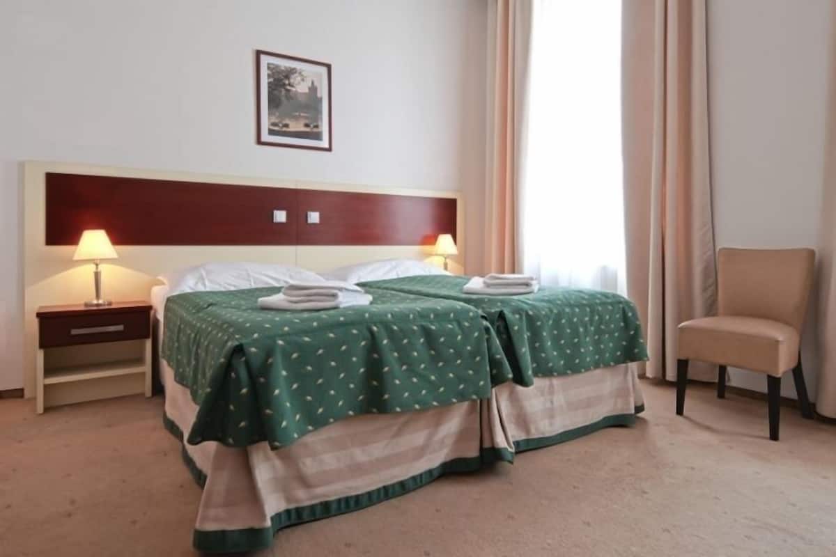 Habitación de hotel con dos camas, una silla, una mesita de noche con lámpara y un cuadro enmarcado en la pared.