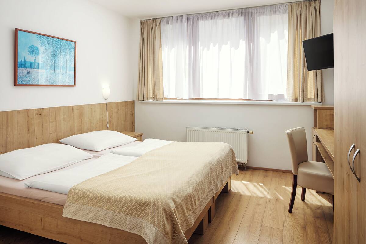A hotel room with a wooden headboard, two beds with white linens, a beige bedspread, a window with sheer curtains, a flat-screen TV mounted above the window, a wooden chair, and a radiator below the window.