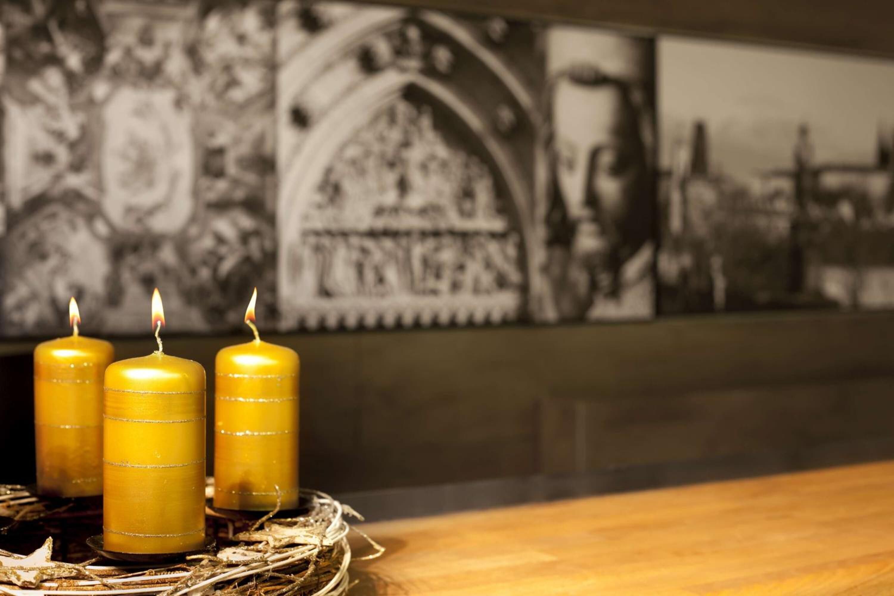Drie brandende kaarsen op een houten oppervlak met een gevlochten mandje eronder, voor een muur met een gotische muurschildering.