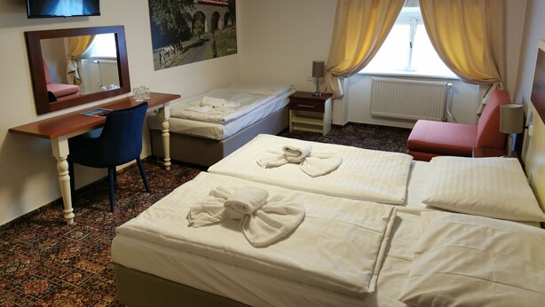 A hotel room with two beds, a desk, a chair, a TV, a window with curtains, and a painting on the wall. - City Inn (Prague)