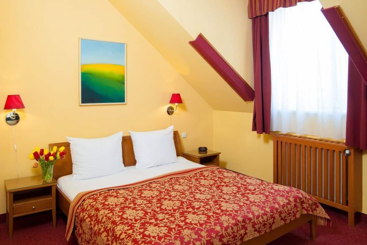 A bedroom with a bed, two bedside tables, a nightstand, a window with curtains, a radiator, and a painting on the wall.