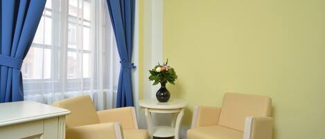 Um quarto com uma escrivaninha, duas cadeiras, uma mesinha com um vaso de flores e uma janela com cortinas azuis.