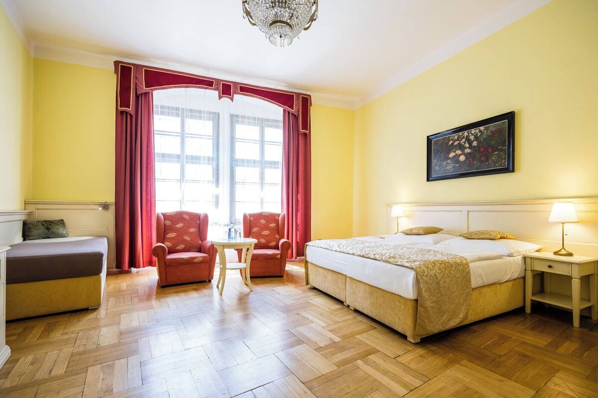 Une chambre d’hôtel comprenant un lit, deux fauteuils, une petite table, une table de chevet, un lustre et un tableau au mur.
