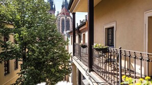 Un edificio con balcón, una iglesia con agujas en el fondo y follaje en primer plano.