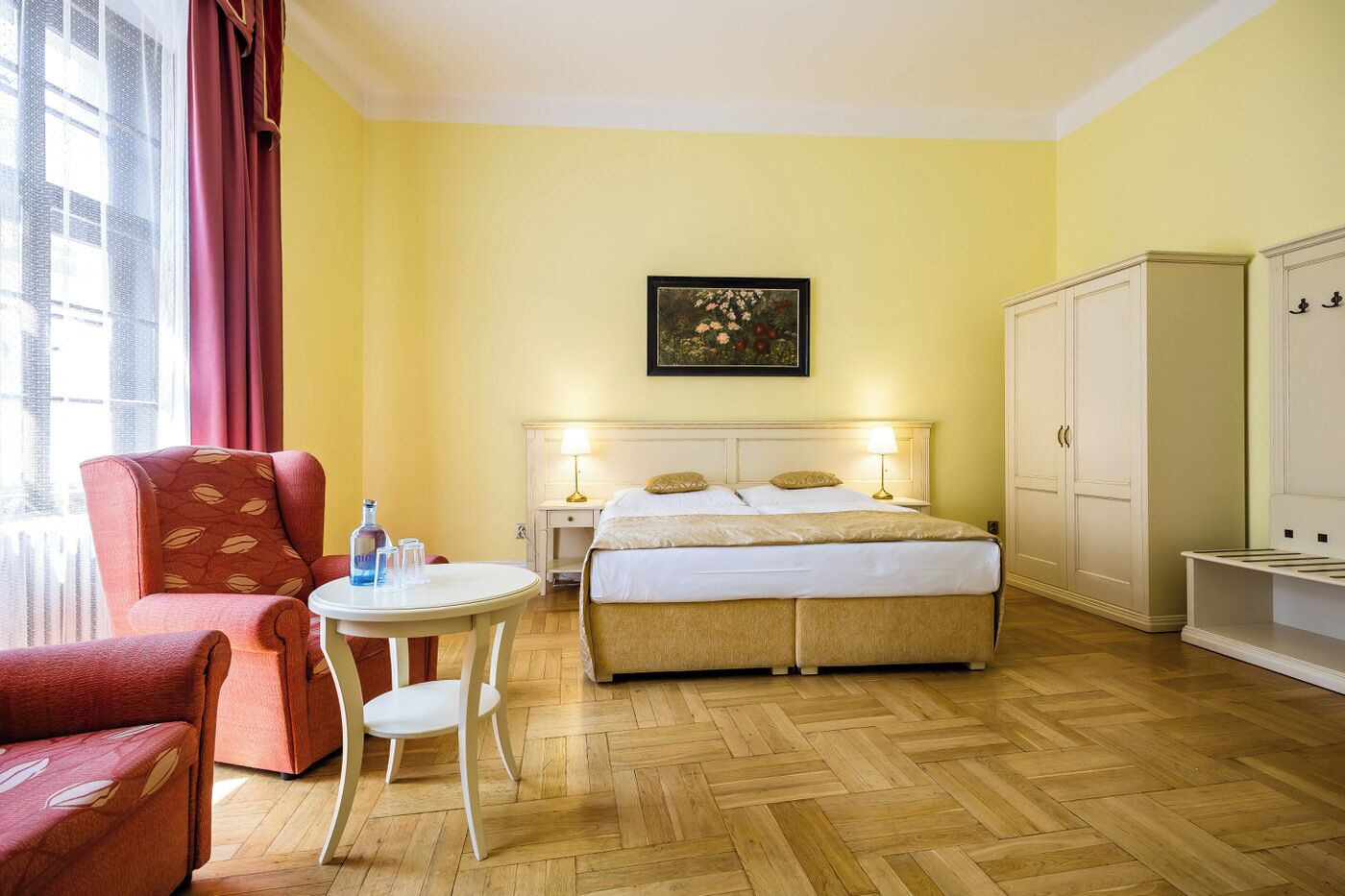 A hotel room with a bed, a chair, a small table, a nightstand, and a wardrobe.