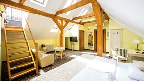 A room with a sloped ceiling, wooden beams, a bed, a desk, a chair, a table, a TV, and a staircase. - Hotel Metamorphis (Prague)
