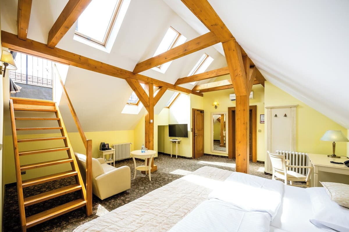 Une pièce avec un plafond en pente, des poutres en bois, un lit, un bureau, une chaise, une table, un téléviseur et un escalier.