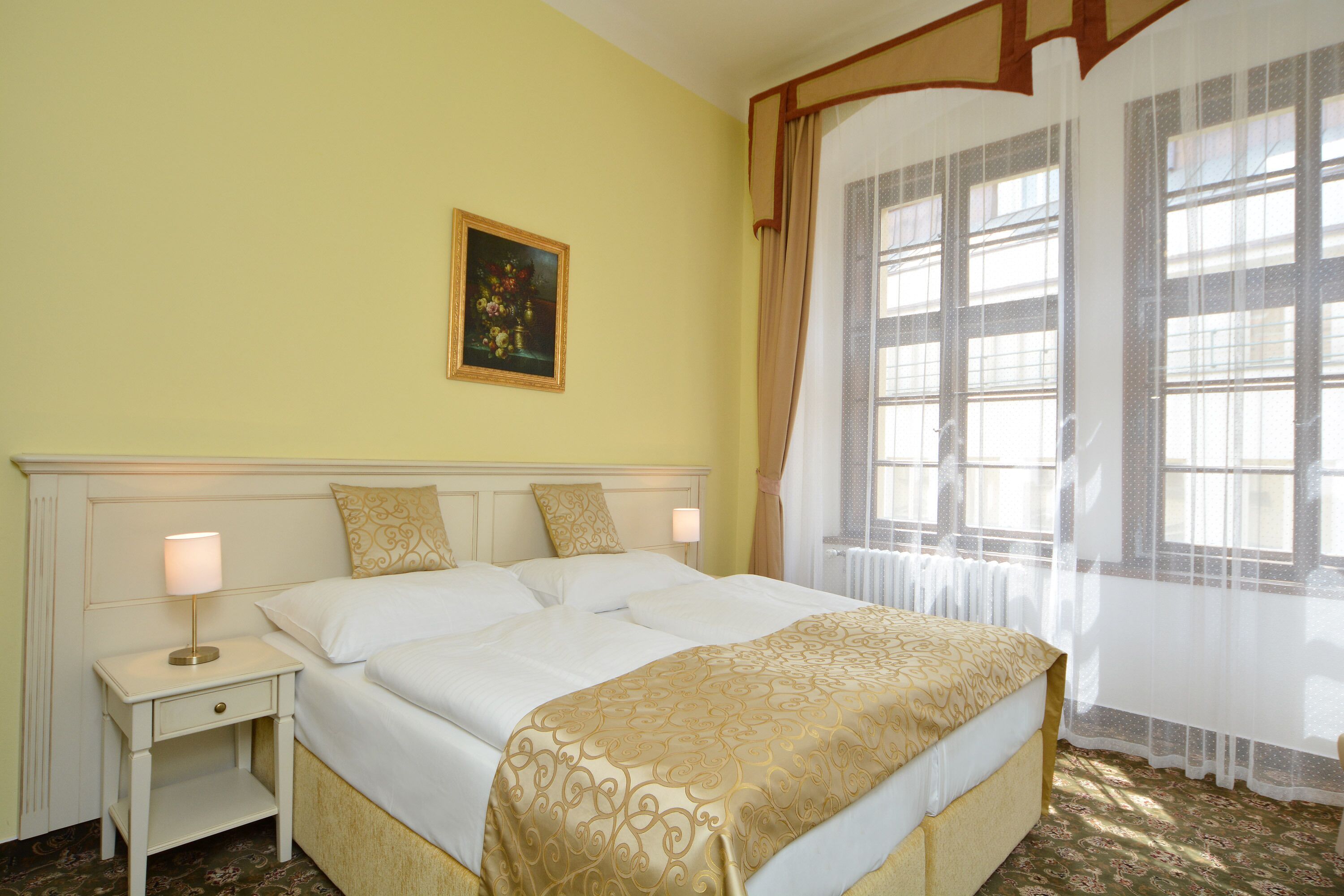 A hotel room with a large bed, a nightstand with a lamp, a painting on the wall, and a window with curtains.