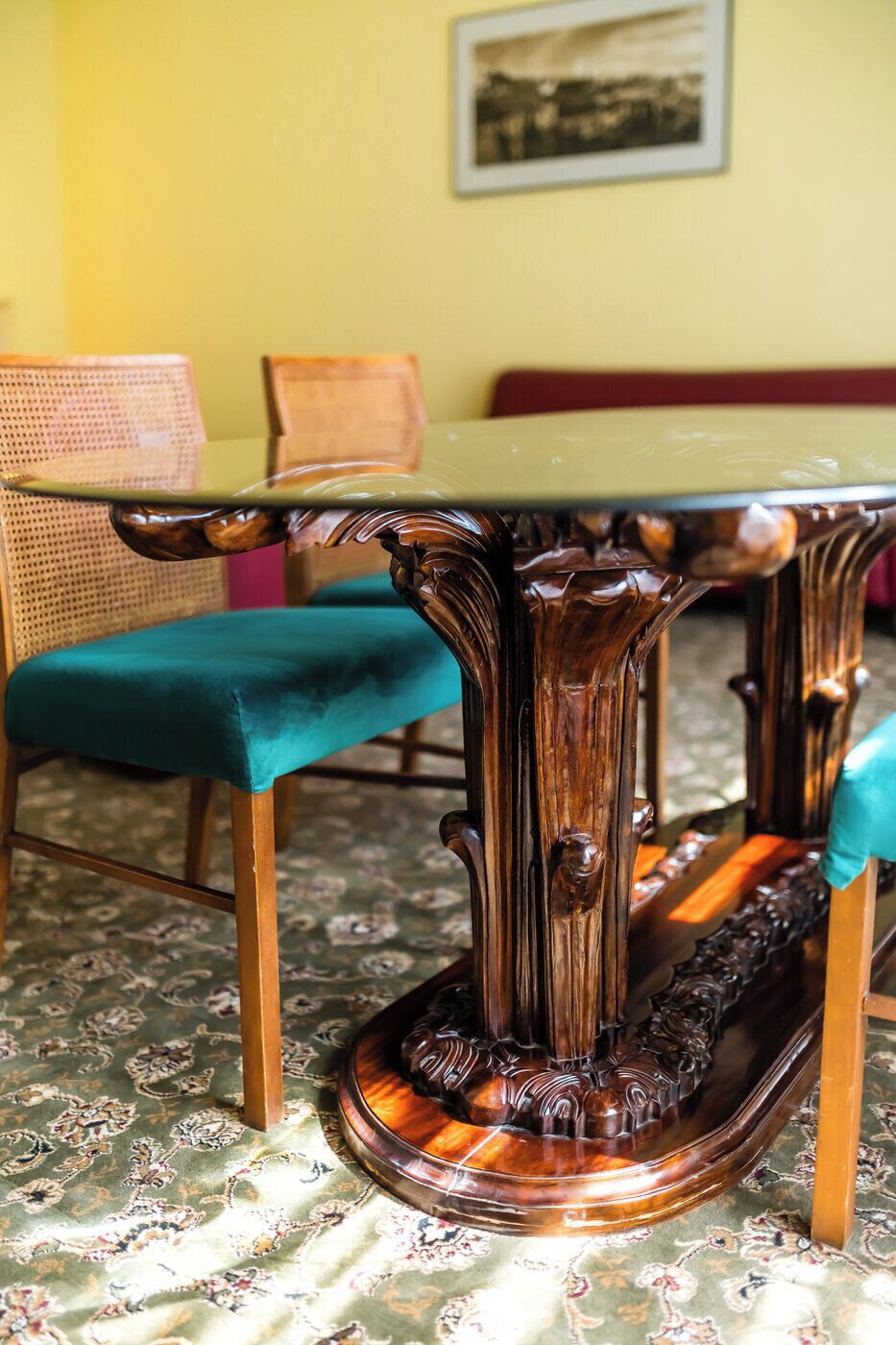 A table with ornate legs and a green top, placed on a patterned carpet in a room with a yellow wall and a framed picture.