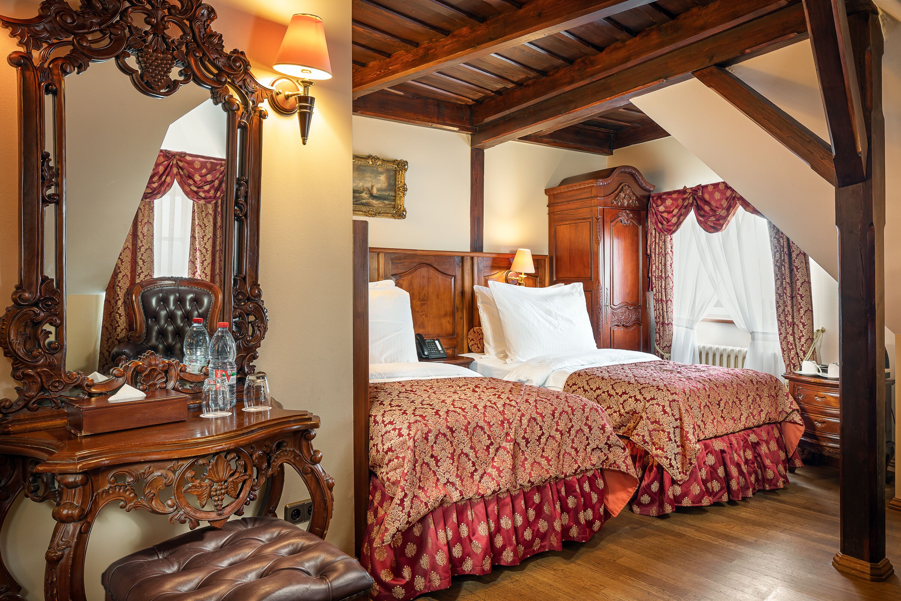 Une chambre à coucher avec un grand miroir décoratif, une tête de lit en bois, deux lits avec une literie à motifs rouges, une commode en bois et un banc.