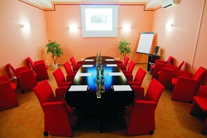 Sala de conferência com uma longa mesa, cadeiras vermelhas, tela de projetor e duas plantas em vasos.