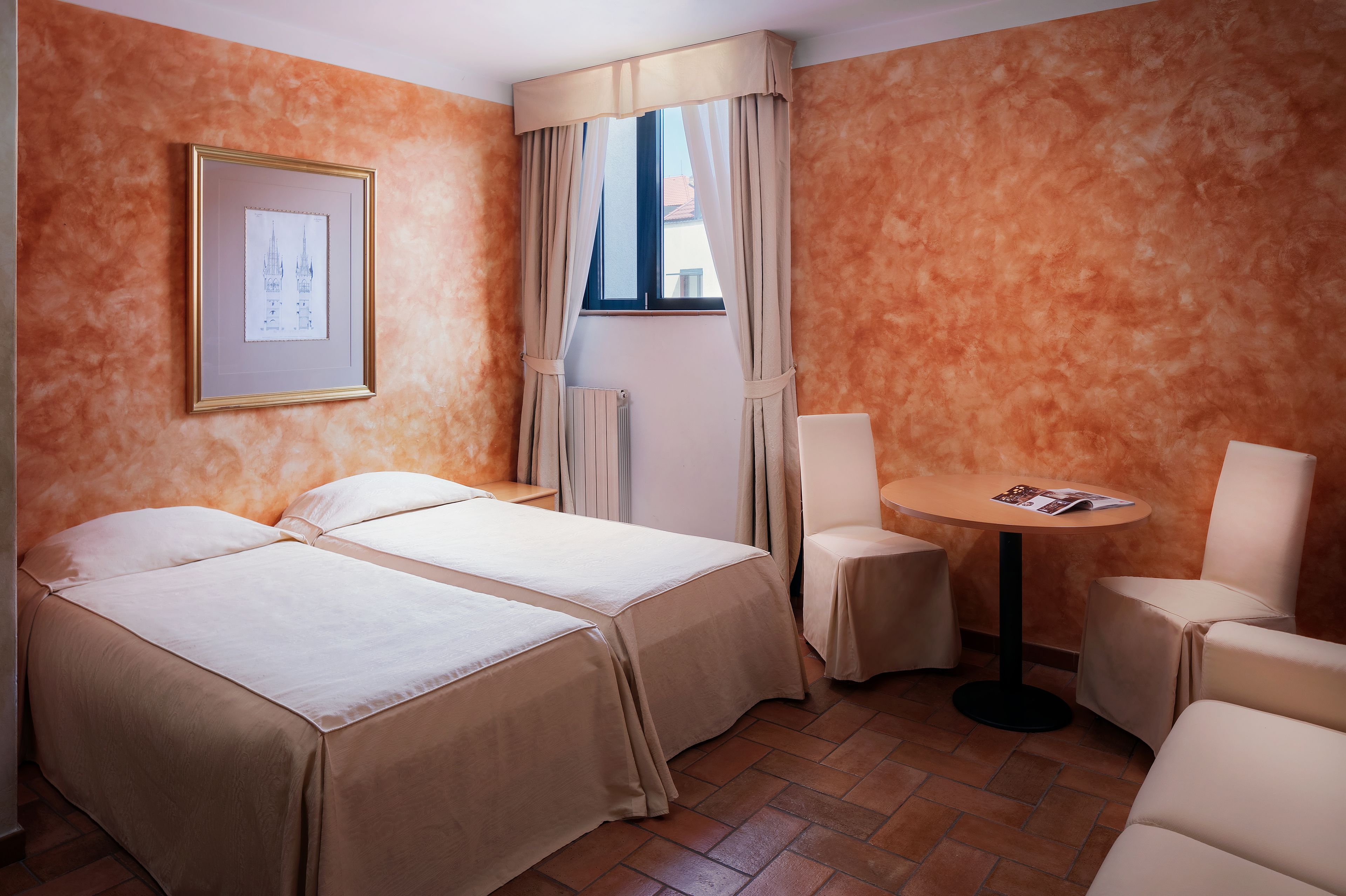 Quarto de hotel com duas camas, uma mesa redonda pequena, uma cadeira e um quadro na parede.