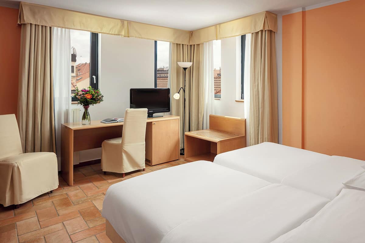 A hotel room with a bed, a desk with a computer, a chair, and a window with curtains.