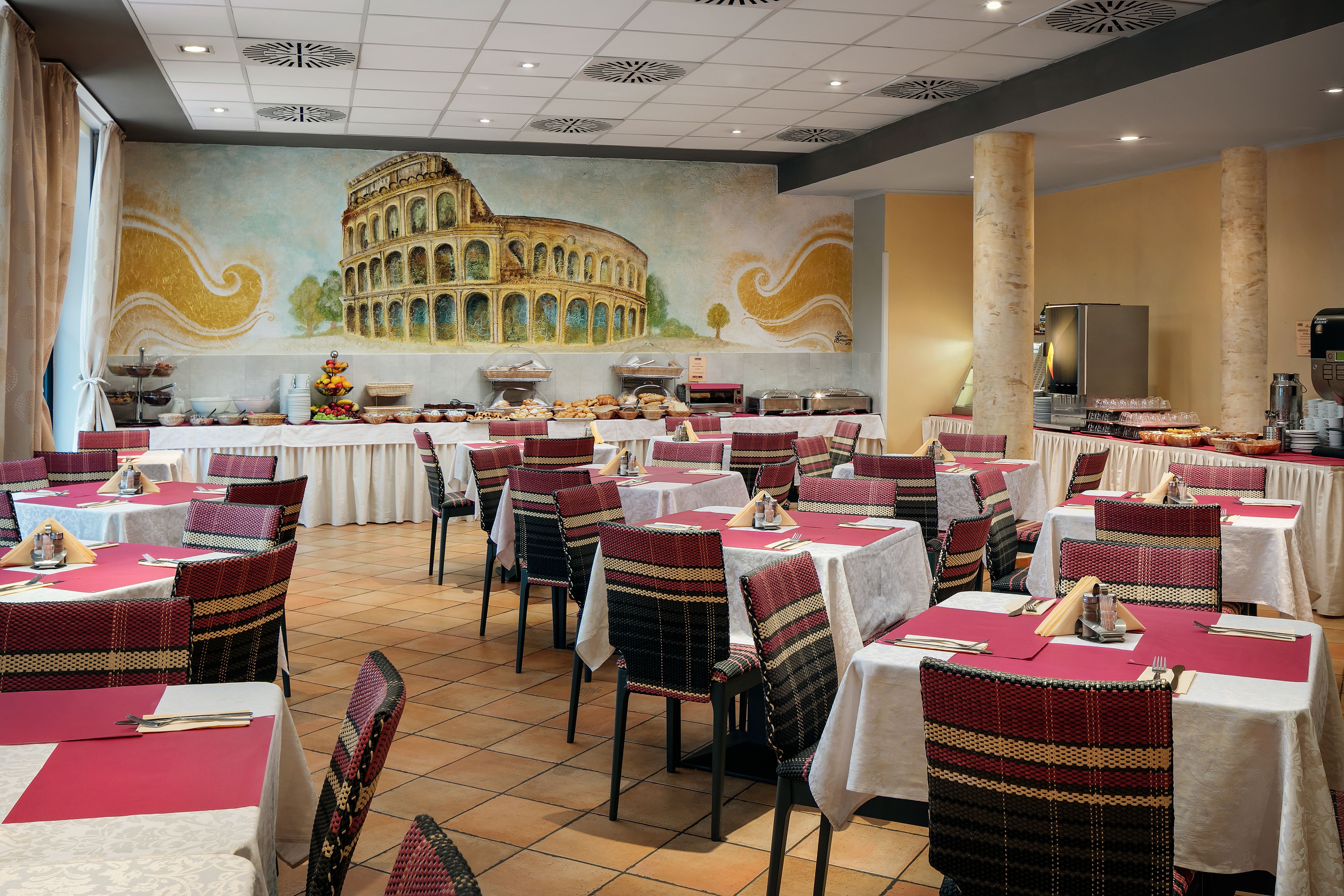 Restaurante com várias mesas e cadeiras, um mural do Coliseu e estações de buffet.