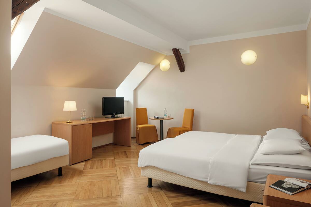 A hotel room with a bed, a desk with a computer, a chair, and a small table with a lamp.