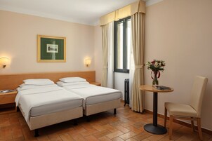 Quarto de hotel com duas camas, uma mesinha com um vaso de flores, uma cadeira e um quadro na parede.