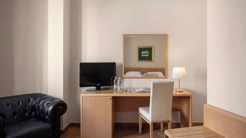 A hotel room with a leather sofa, a wooden desk with a computer, a chair, a bedside table with a lamp, and a bed with a headboard.