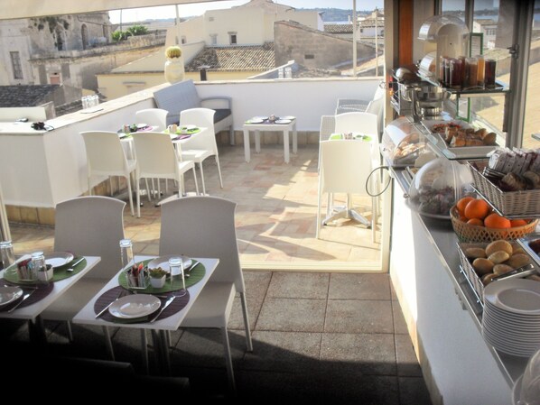 A rooftop dining area with tables and chairs, a buffet with various food items, and a view of the cityscape.