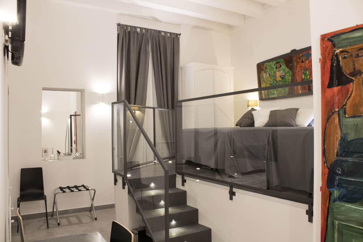 Un dormitorio moderno con una escalera de cristal, una cama con ropa de cama gris, un cuadro en la pared y una mesita de noche con una silla.