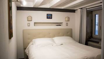 Een net opgemaakt bed in een kleine kamer met een hoofdeinde, een plank boven het bed en een raam met gordijnen.