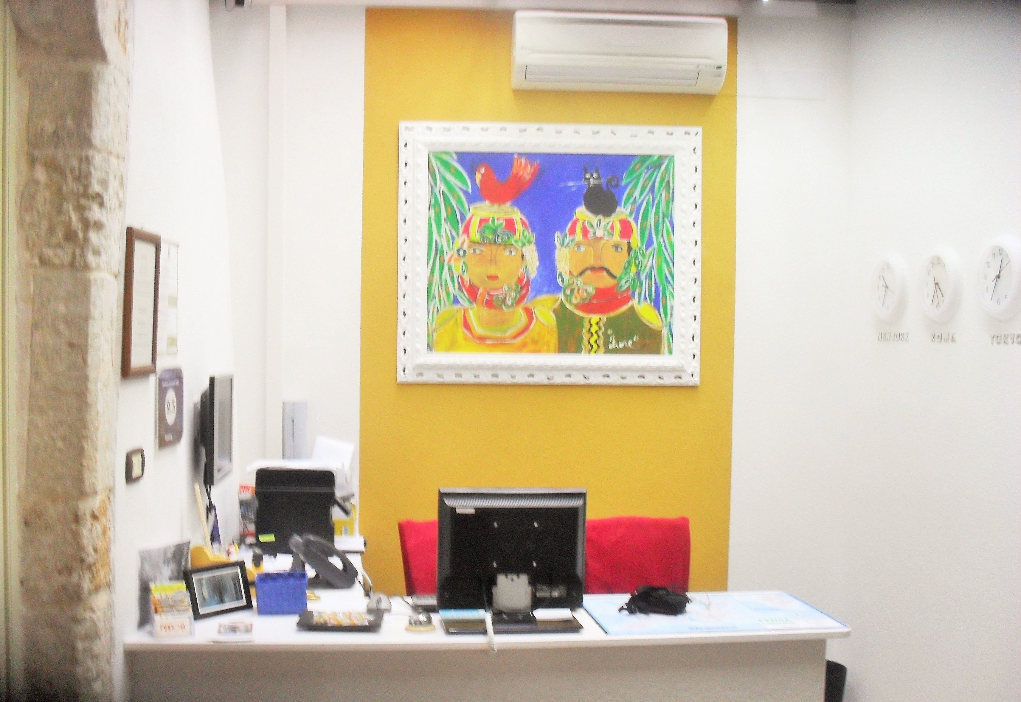 A reception desk with a computer, phone, and various office supplies. There is a framed painting above the desk and a clock on the wall to the right.