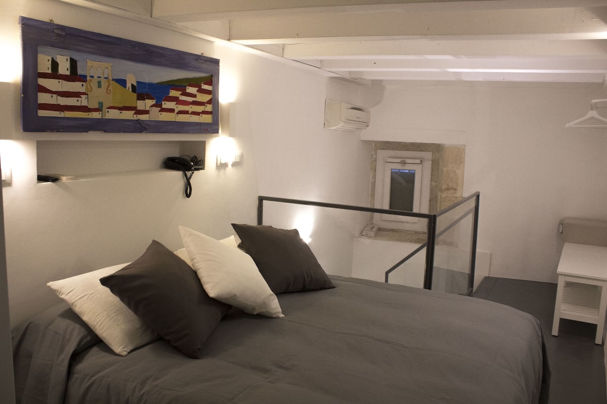 Un dormitorio con cama, almohadas, un cuadro, un teléfono y una mesita.