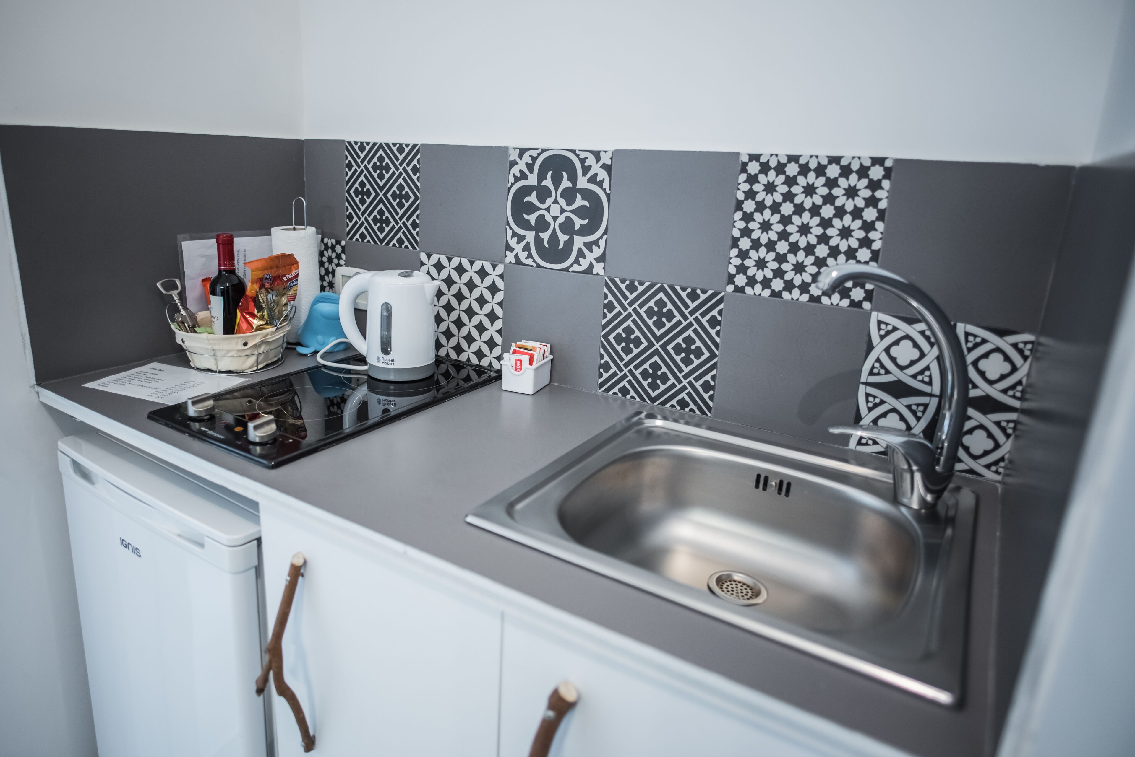 A kitchen with a stainless steel sink, a kettle, a basket with utensils, and a coffee maker.