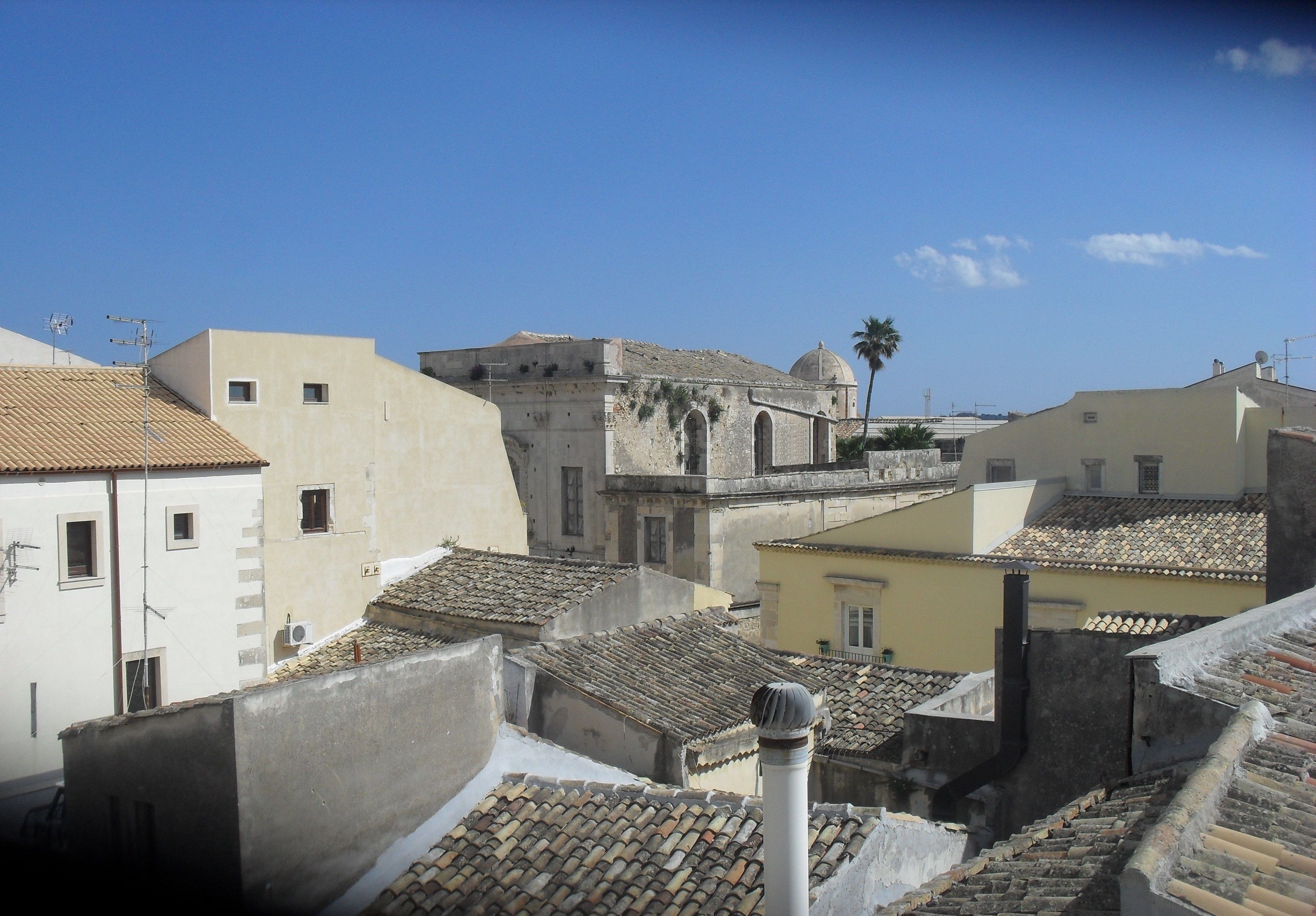 A view of a historic town with terracotta-tiled rooftops, a prominent building with a dome, and a palm tree in the distance.