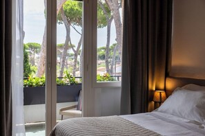 A hotel room with a bed, a nightstand, a lamp, and a view of trees and buildings through large windows. - Hotel le Pleiadi (Forte dei Marmi)