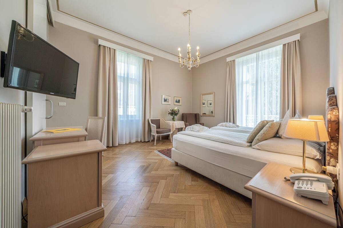 A hotel room with a bed, a desk, a chair, a TV, and a chandelier.