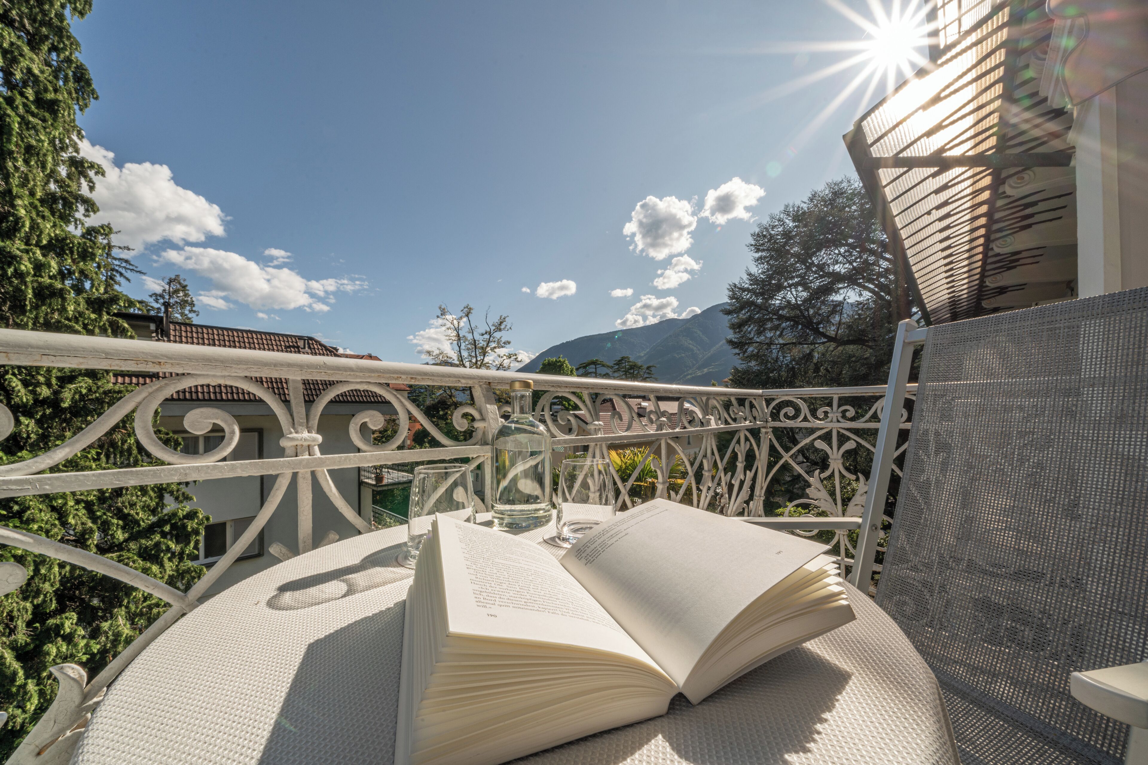 An open book on a round table with a metal railing and a mesh chair, set against a backdrop of trees and a mountain.