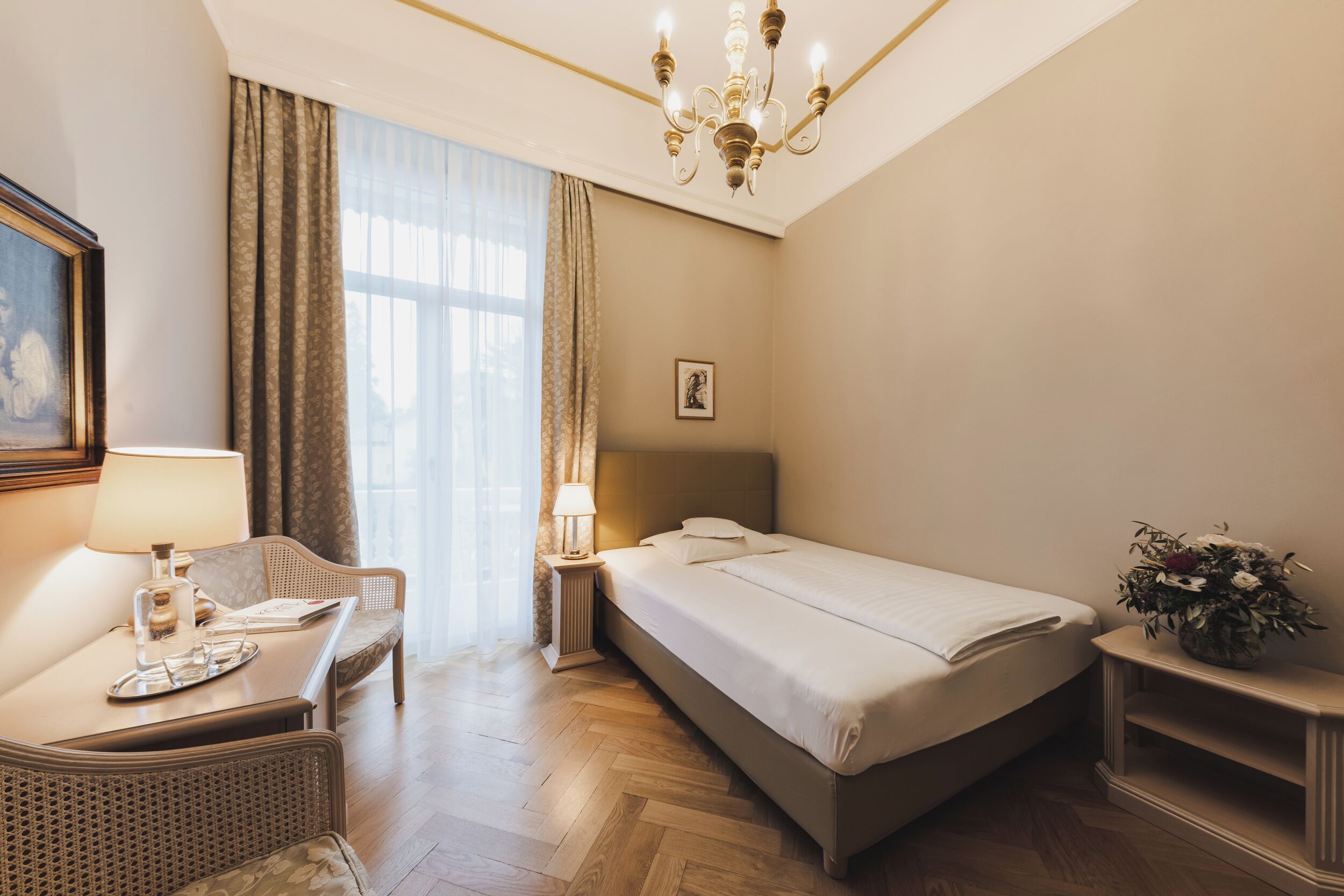 A hotel room with a bed, a chair, a nightstand with a lamp, a picture on the wall, and a window with curtains.