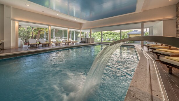 Une piscine intérieure moderne agrémentée d’une cascade, de chaises longues et de grandes fenêtres offrant une vue sur l’extérieur.
