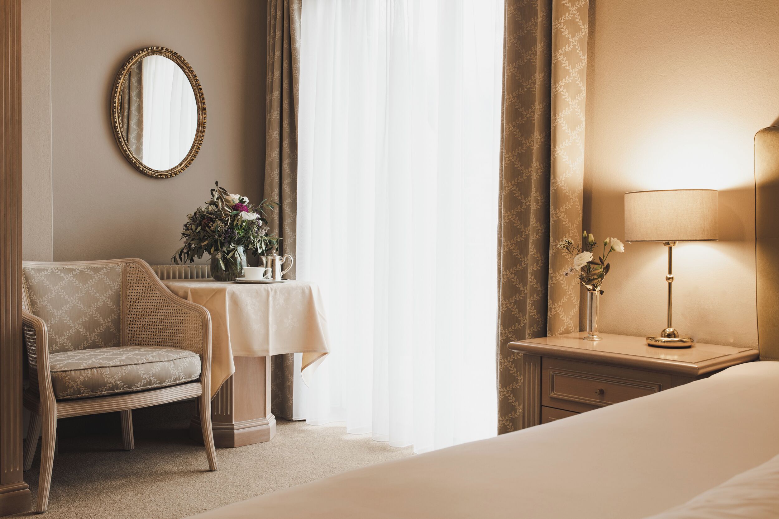 A hotel room with a bed, a bedside table with a lamp, a chair, a table with a vase of flowers, and a mirror on the wall.
