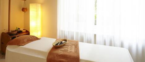 A hotel room with a bed, a massage table, a lamp, and a window with curtains.