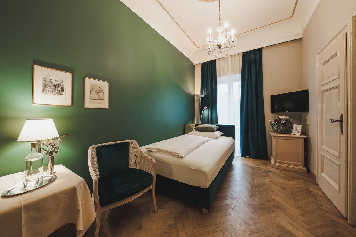A hotel room with a bed, a chair, a table with a lamp, a TV, and a door.