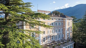 Un hôtel à plusieurs étages doté d’un balcon et d’un espace extérieur aménagé, entouré d’une végétation luxuriante et avec une vue sur les montagnes en arrière-plan.