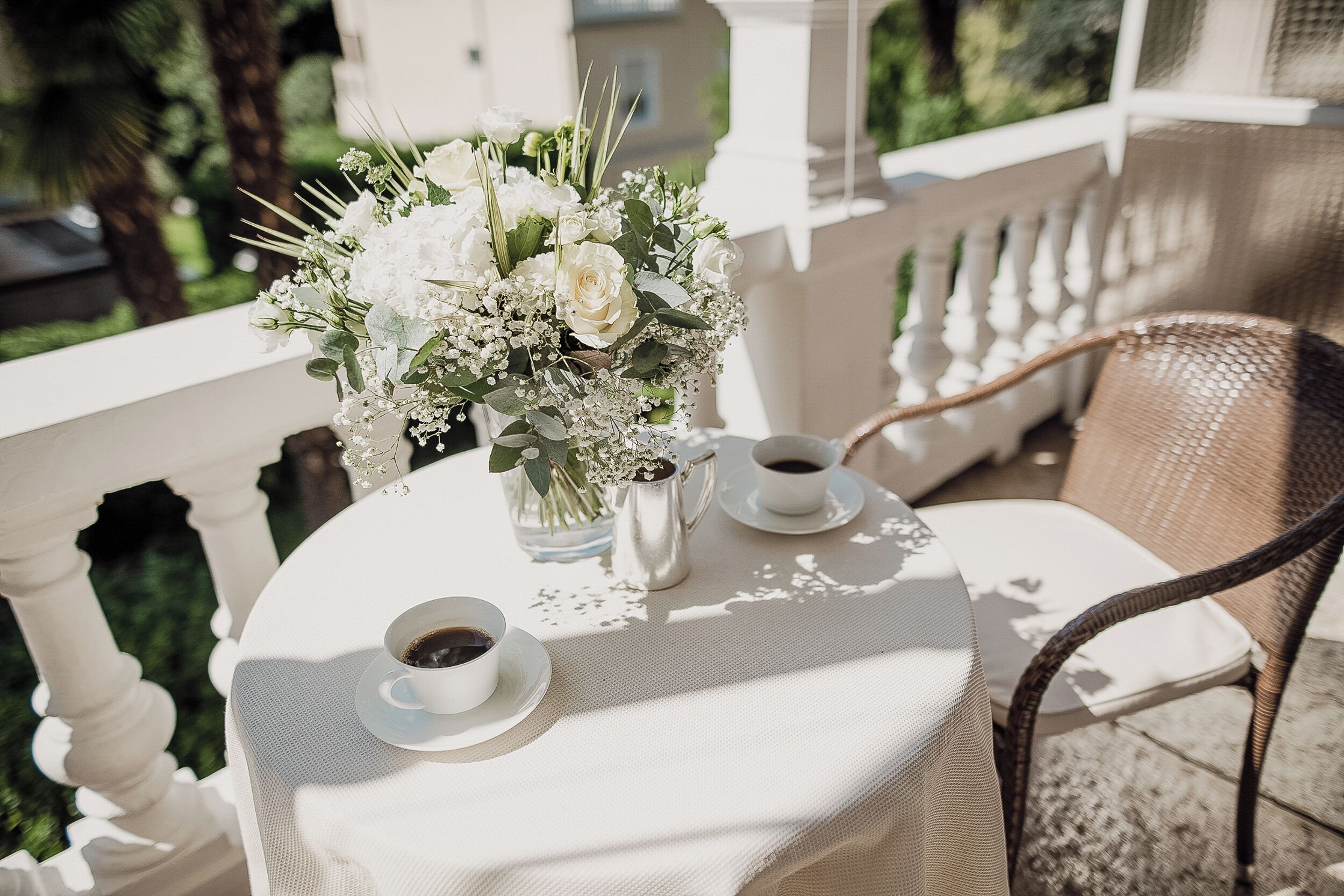 A round table with a white tablecloth, two white cups of coffee, a silver pitcher, and a bouquet of white flowers.