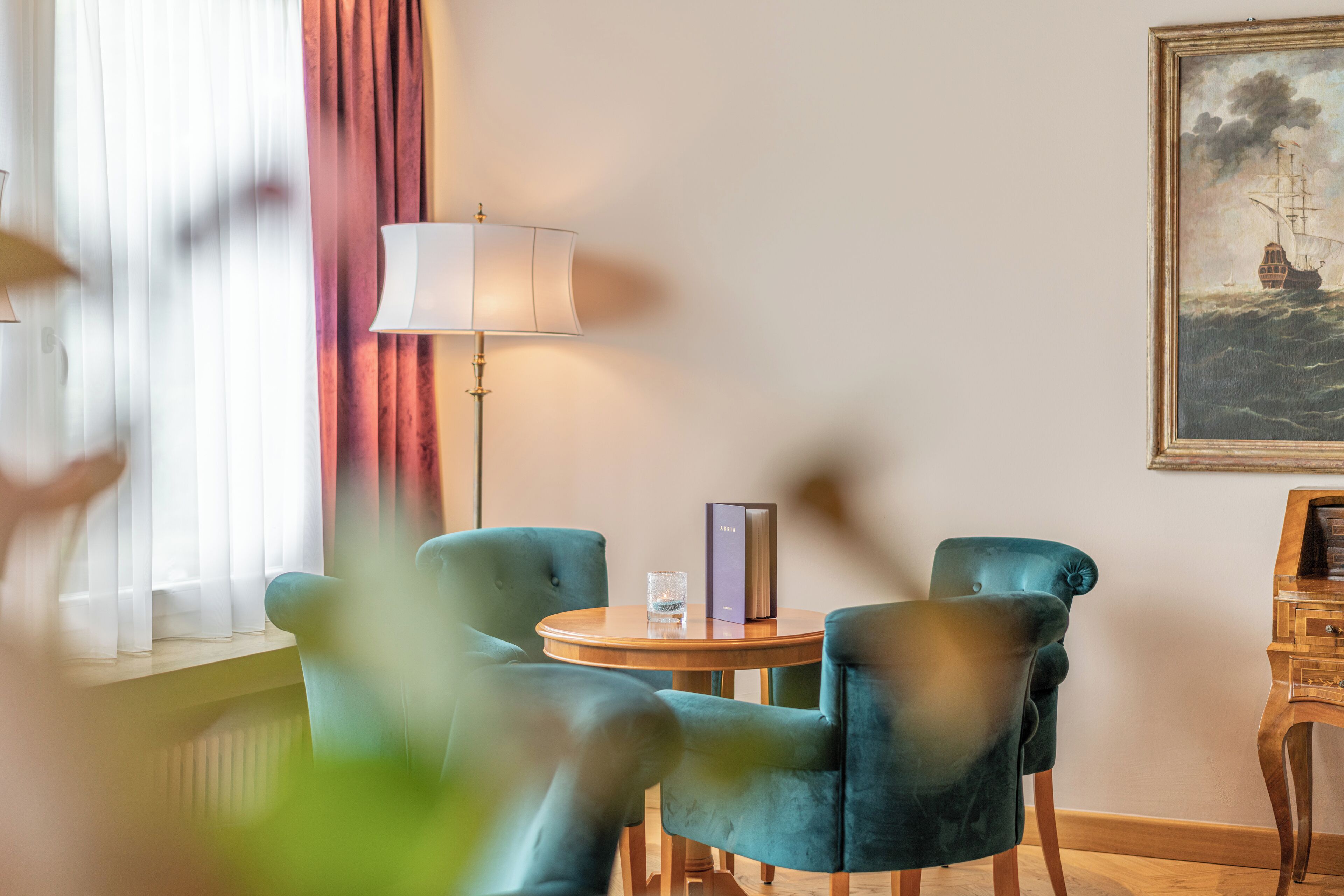 A room with two teal chairs, a wooden table, a lamp, and a framed painting on the wall.