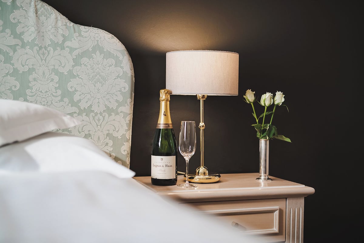 A neatly made bed with a pillow, a bedside table with a bottle of champagne, a glass, and a vase with flowers.