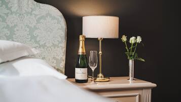 Una cama bien hecha con un almohadón, una mesita de noche con una botella de champán, una copa y un florero con flores.