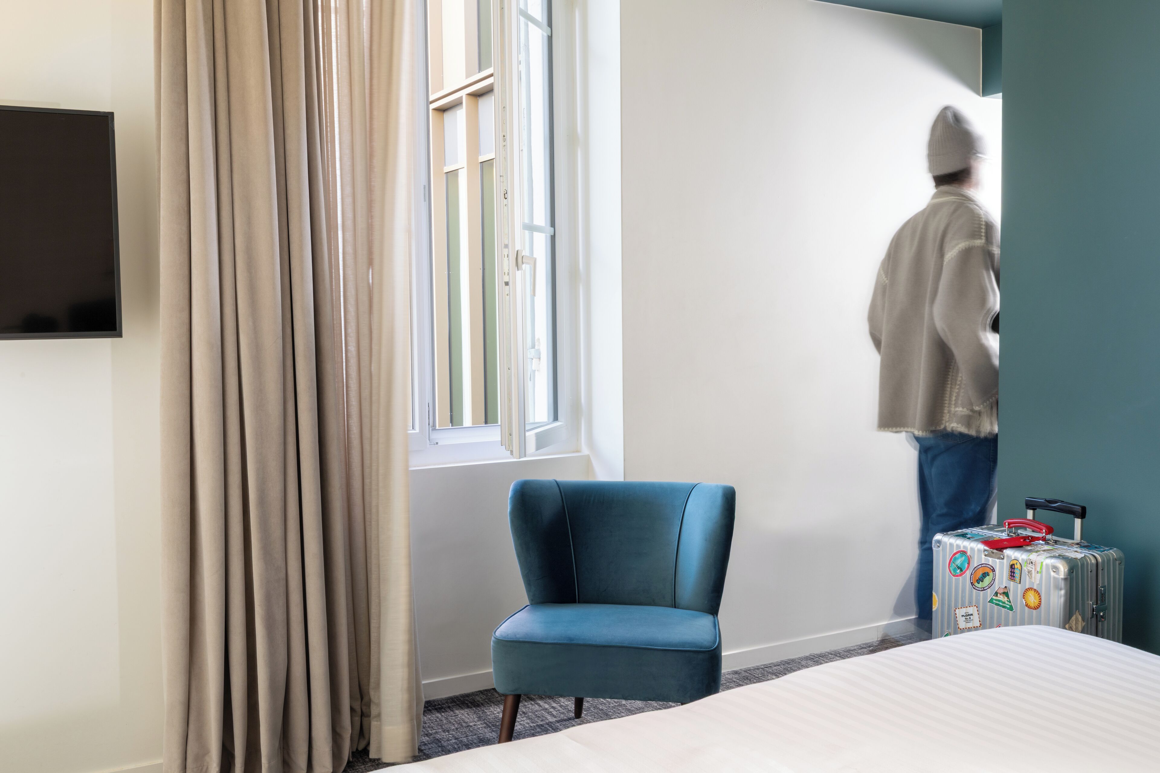 Hotelkamer met een bed, een blauwe fauteuil, een raam met gordijnen, een televisie aan de muur en een koffer met stickers.
