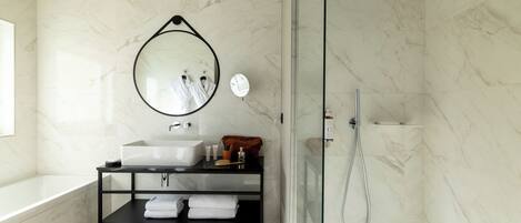 Een moderne badkamer met een douche in een glazen cabine, een wand betegeld met marmer, een ronde spiegel en een zwart plankje met handdoeken.
