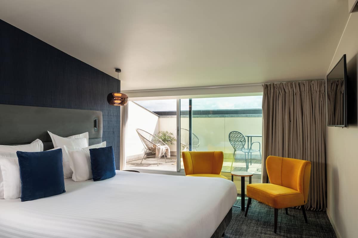 A hotel room with a large bed, a sofa, a yellow armchair, a small table, and a balcony with outdoor seating.
