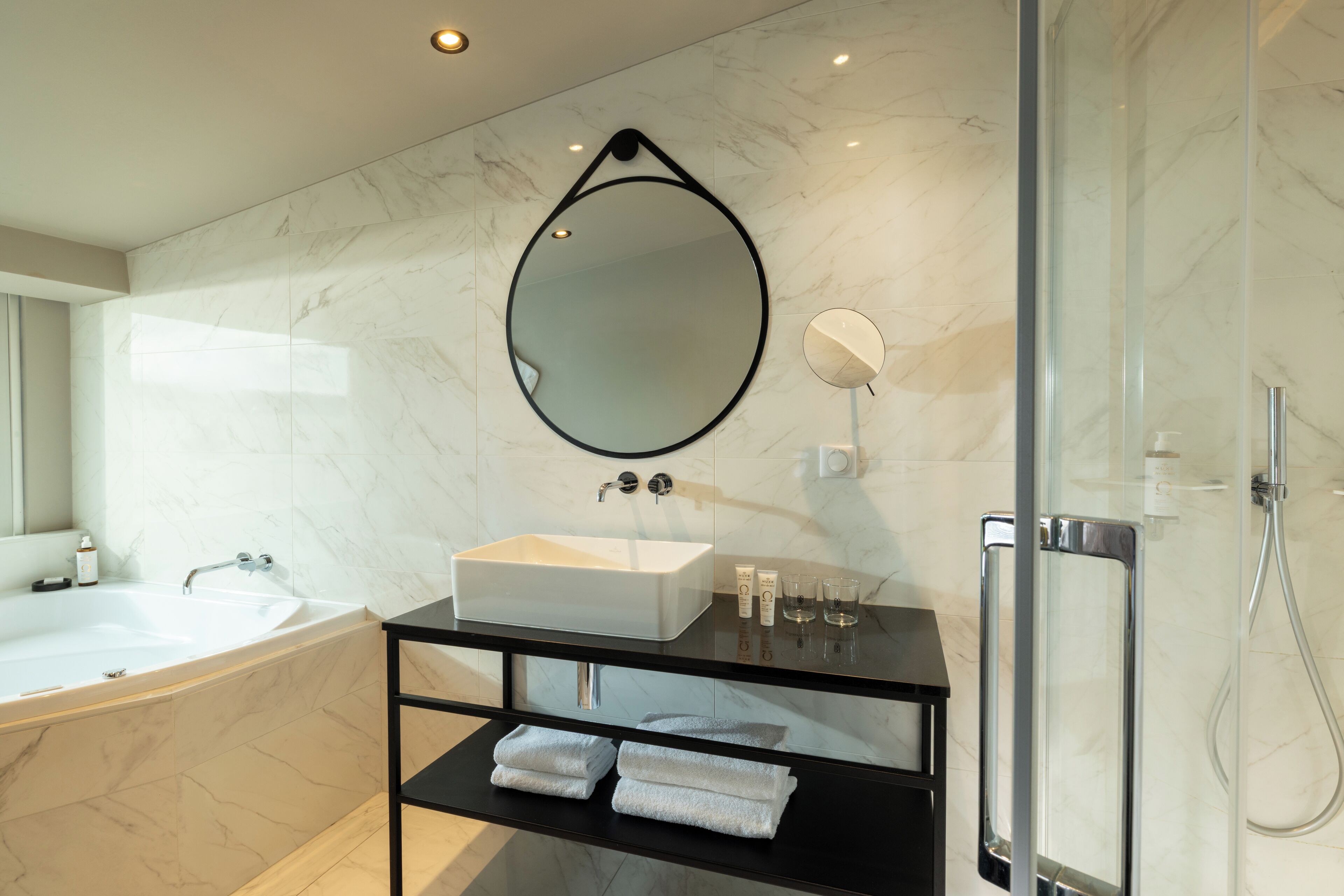 A modern bathroom with a white bathtub, a large mirror, a glass-enclosed shower, and neatly folded towels on a black shelf.