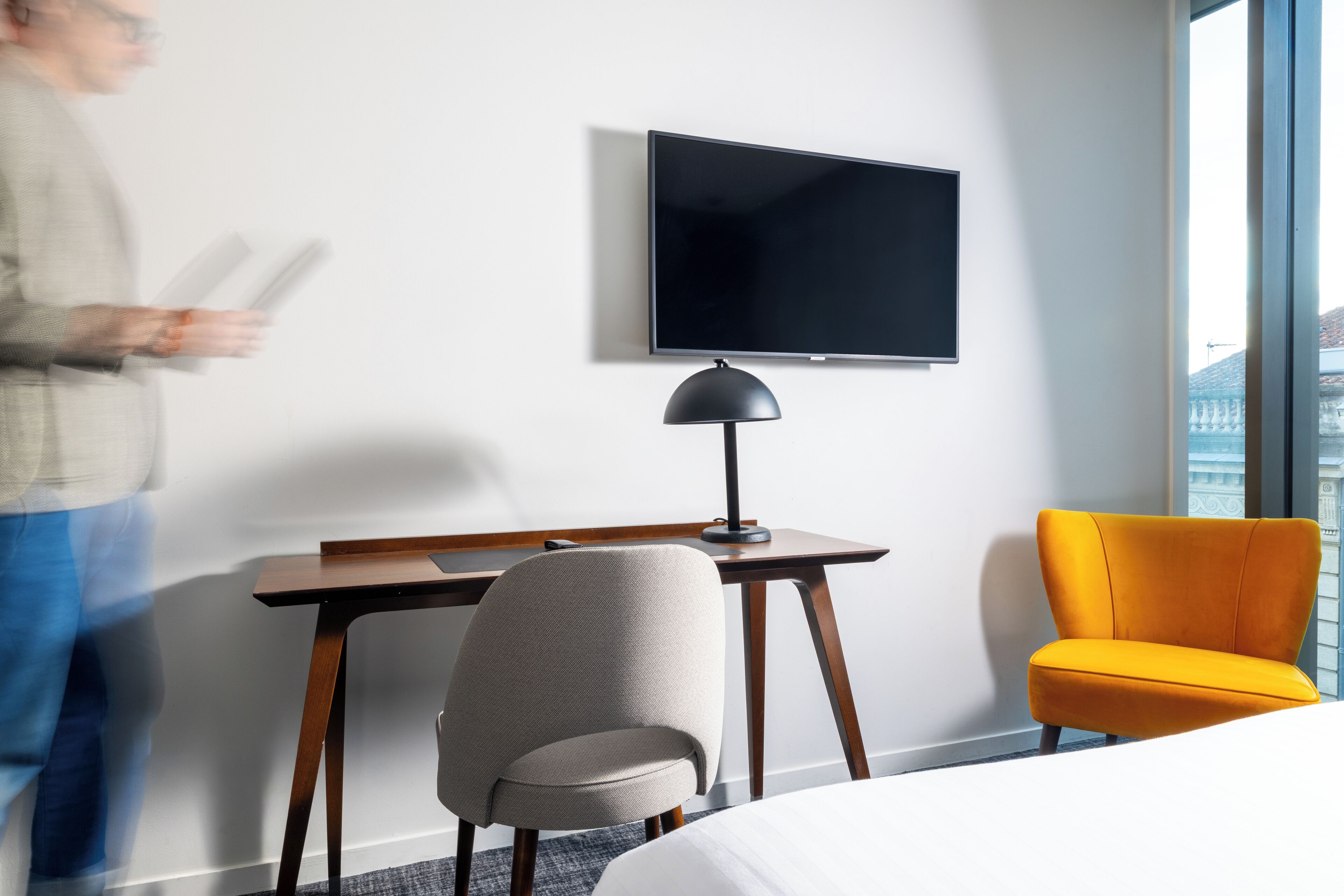 Une chambre d’hôtel comprenant un lit, un bureau avec une chaise, une télévision et une fenêtre donnant sur des immeubles.