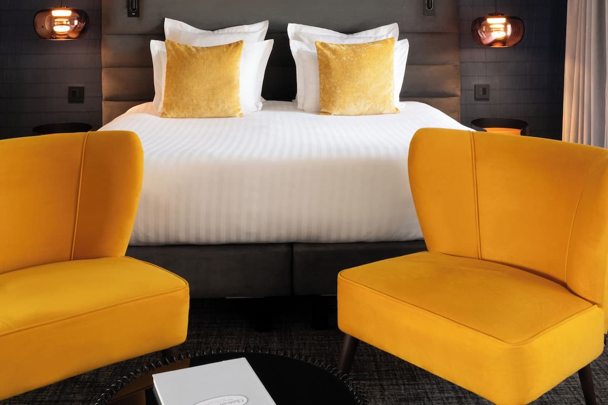 A hotel room with a large bed, two yellow armchairs, a small table, and a book on the table.