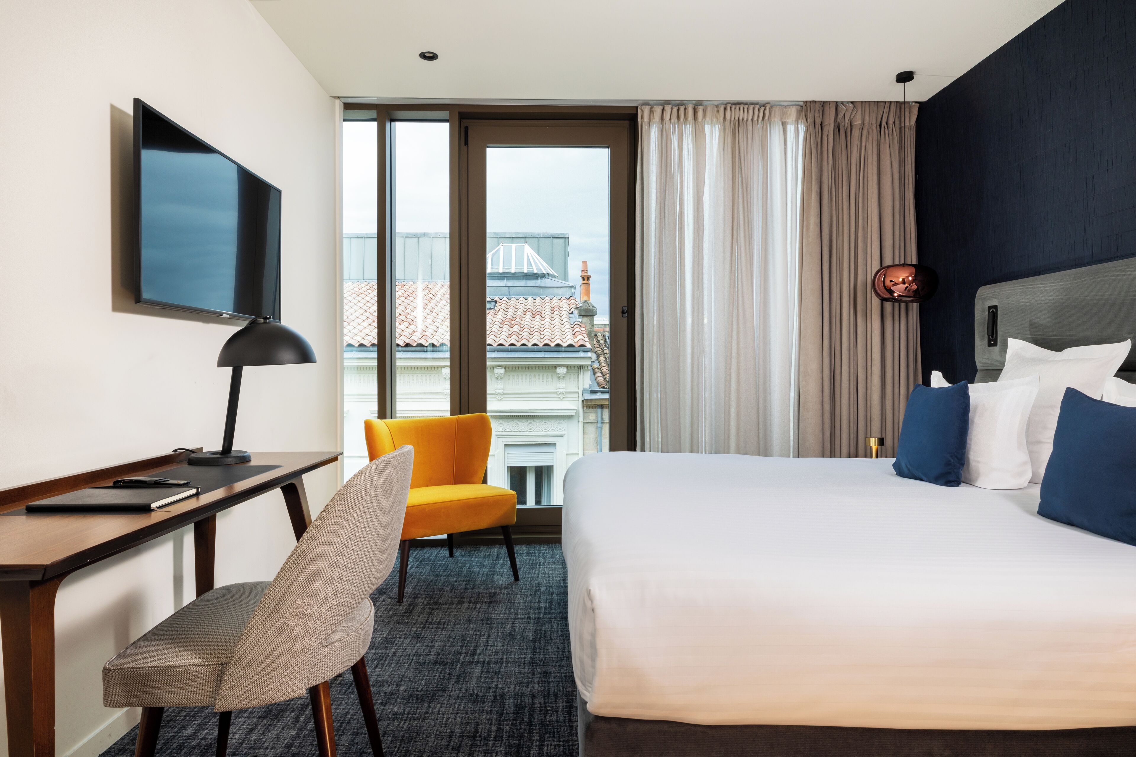 Ein Hotelzimmer mit Bett, Schreibtisch mit Stuhl, Fernseher und Blick auf Gebäude durch das Fenster.