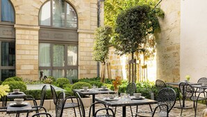 An outdoor patio with tables and chairs, surrounded by greenery and a stone building with arched windows. - La Maison Bord'eaux (Bordeaux)