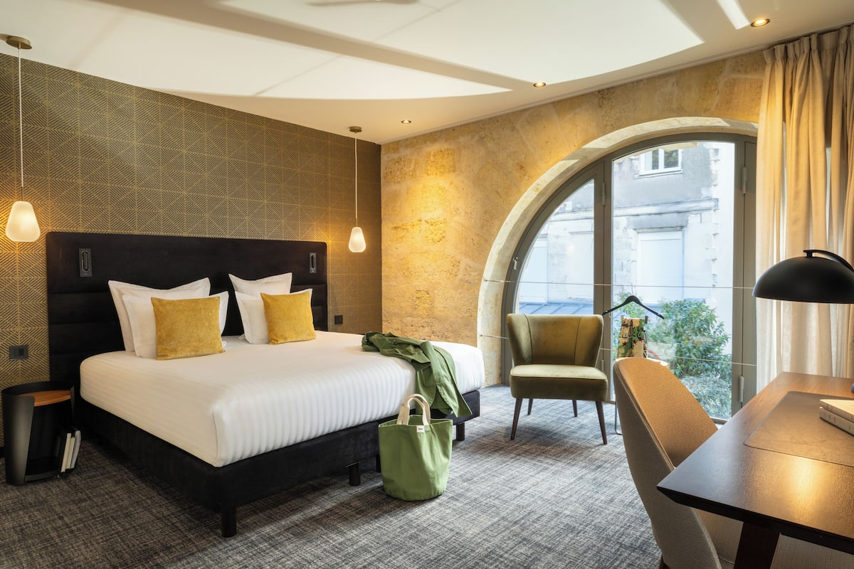 A hotel room with a large bed, a desk, a chair, and a window with a view of buildings.